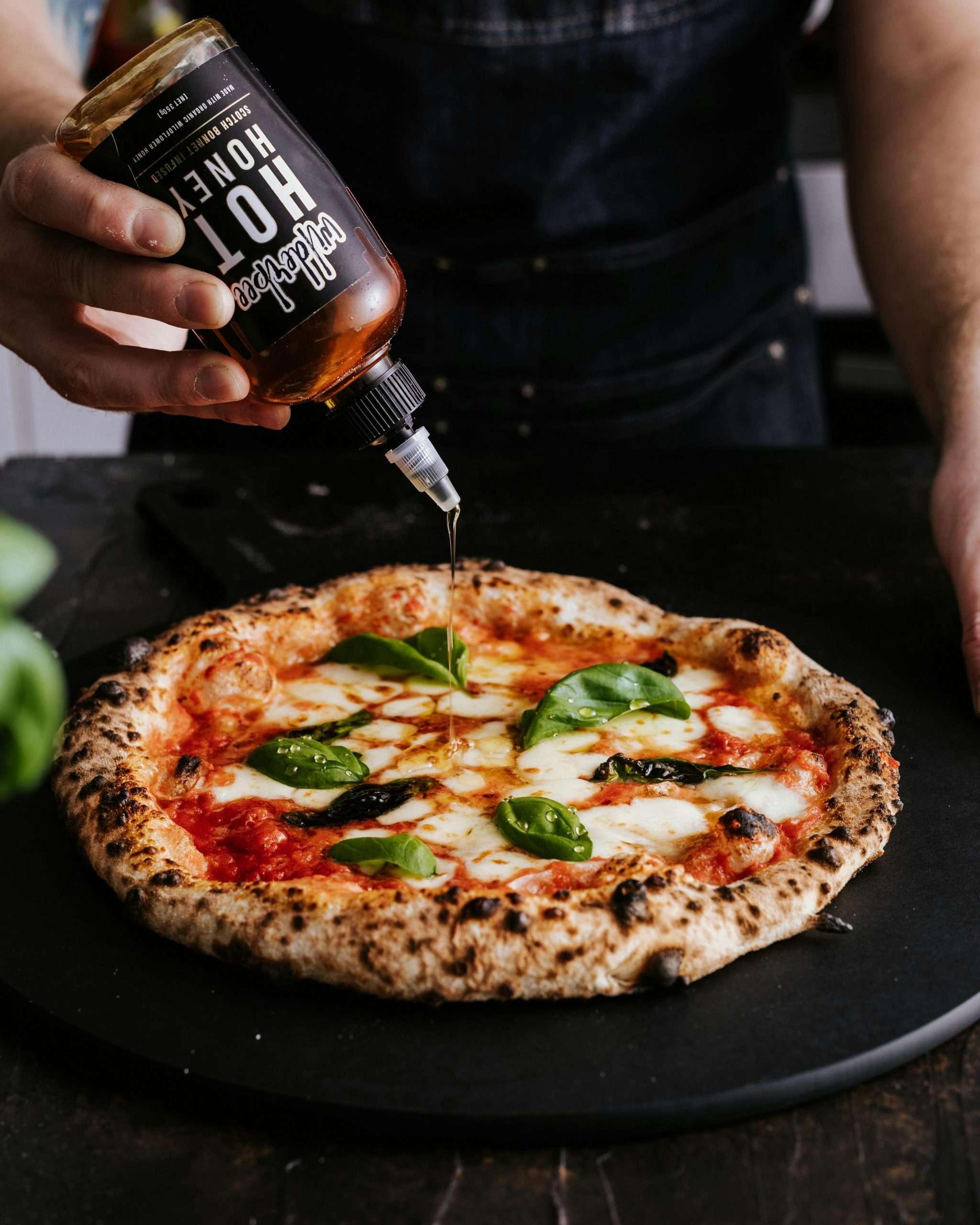 Close-up of a sourdough pizza with basil and mozzarella being drizzled with spicy honey.